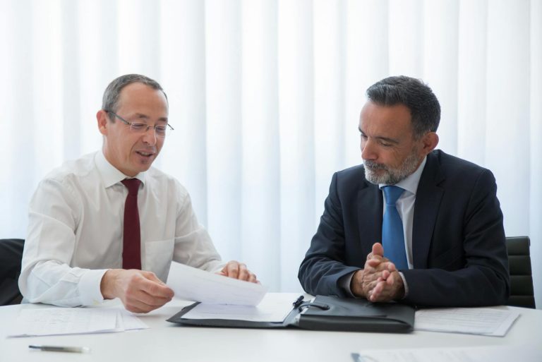 Two businessmen in suits having a discussion over documents in a bright office setting.